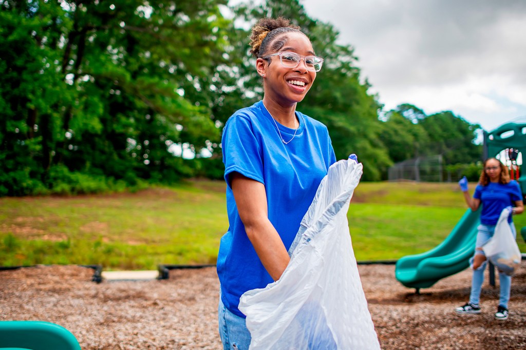 A person stands outdoors holding a large bag while helping clean up a park, with another person picking up trash in the background near a playground.