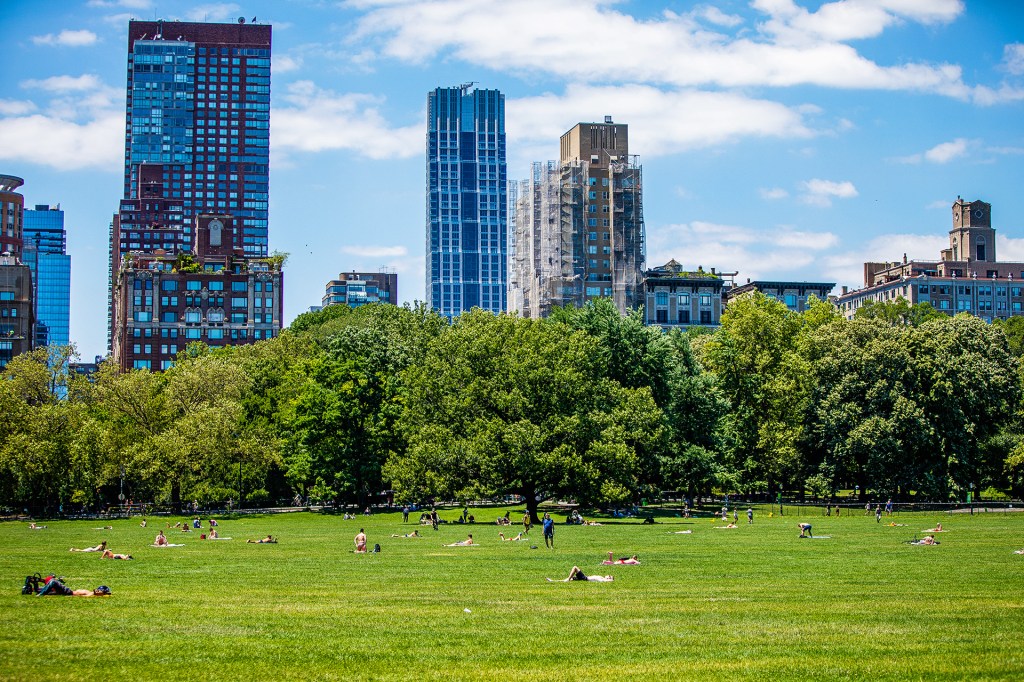 A city skyline rises behind a large green park where people are sitting and lying on the grass.