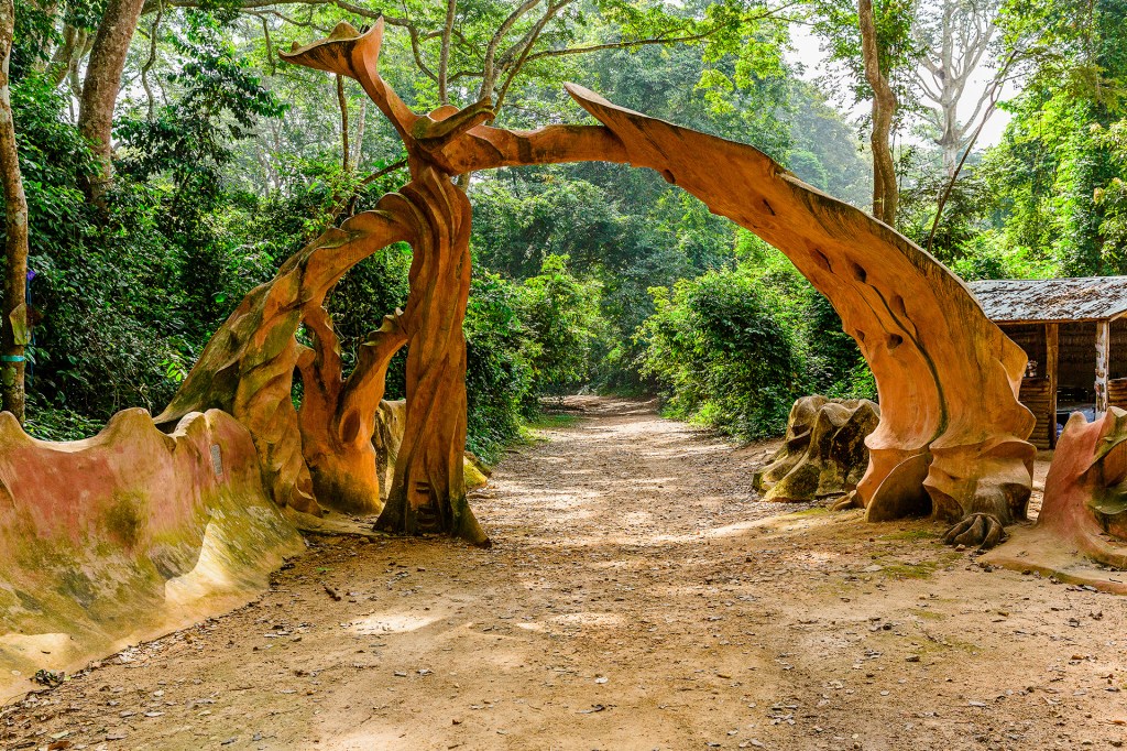A curved wooden arch sculpture stands over a dirt path in a forest surrounded by tall trees and greenery.