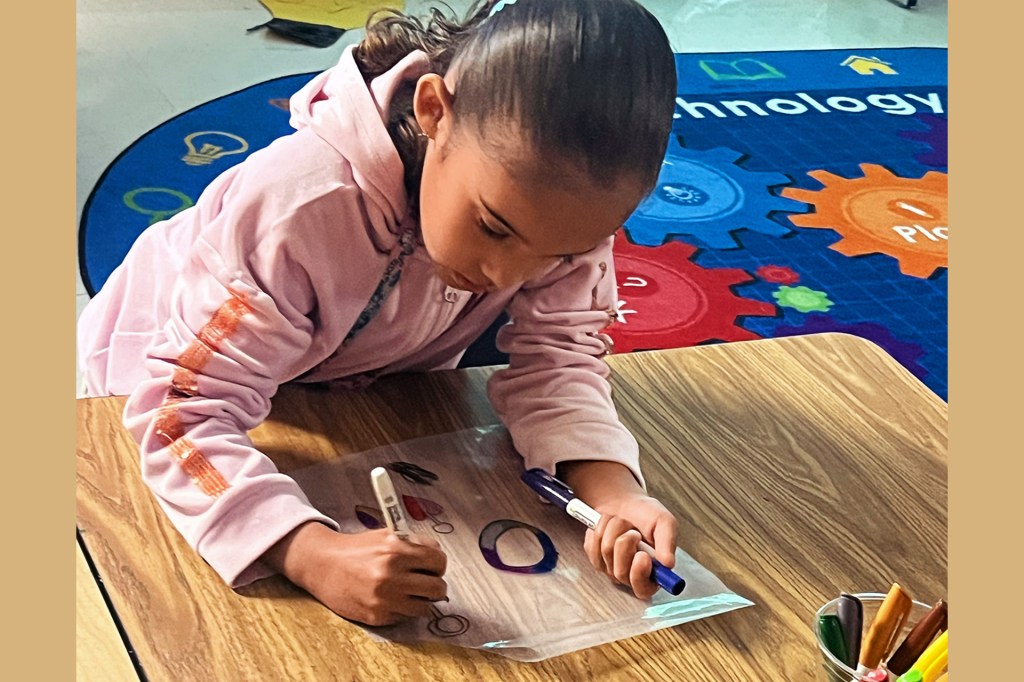 A girl draws with markers on paper at a desk.