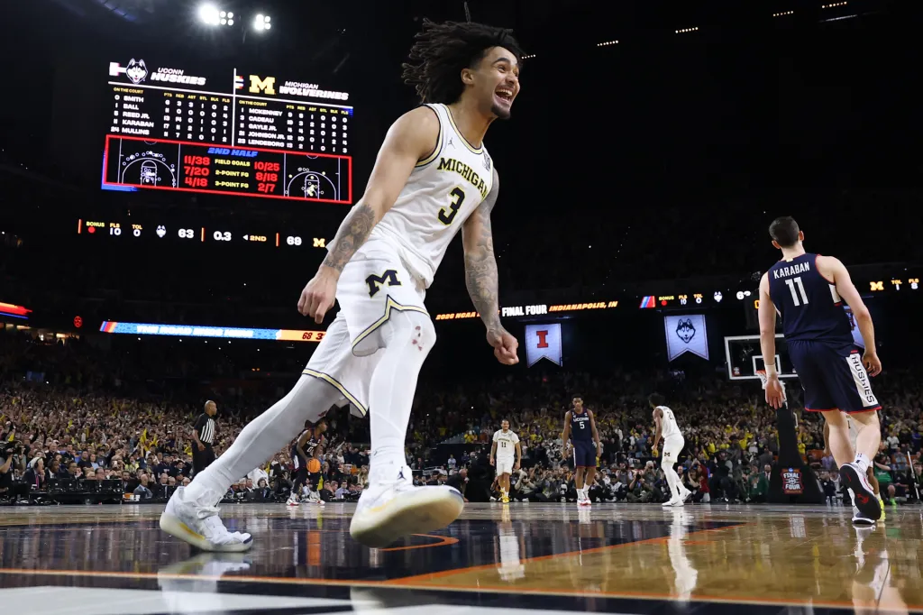 A basketball player in a white Michigan uniform celebrates on the court during a game, smiling as teammates and opponents move in the background and a scoreboard glows overhead.