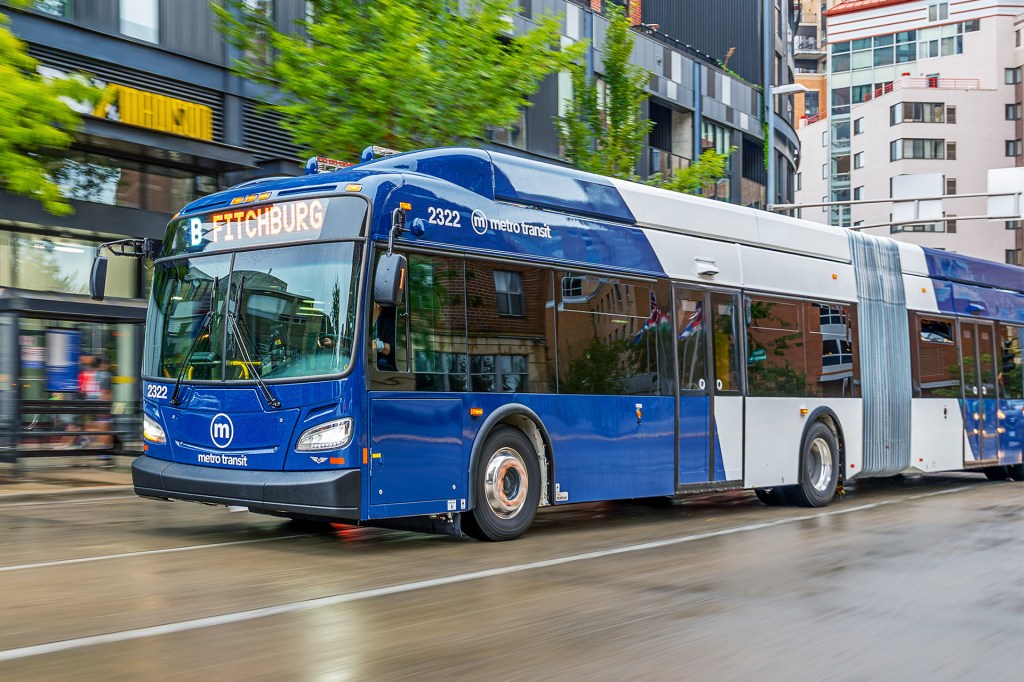A long blue city bus drives down a wet street in a busy urban area.