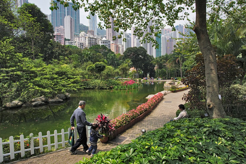 A man and a child walk along a path beside a pond in a green city park with tall buildings in the background.