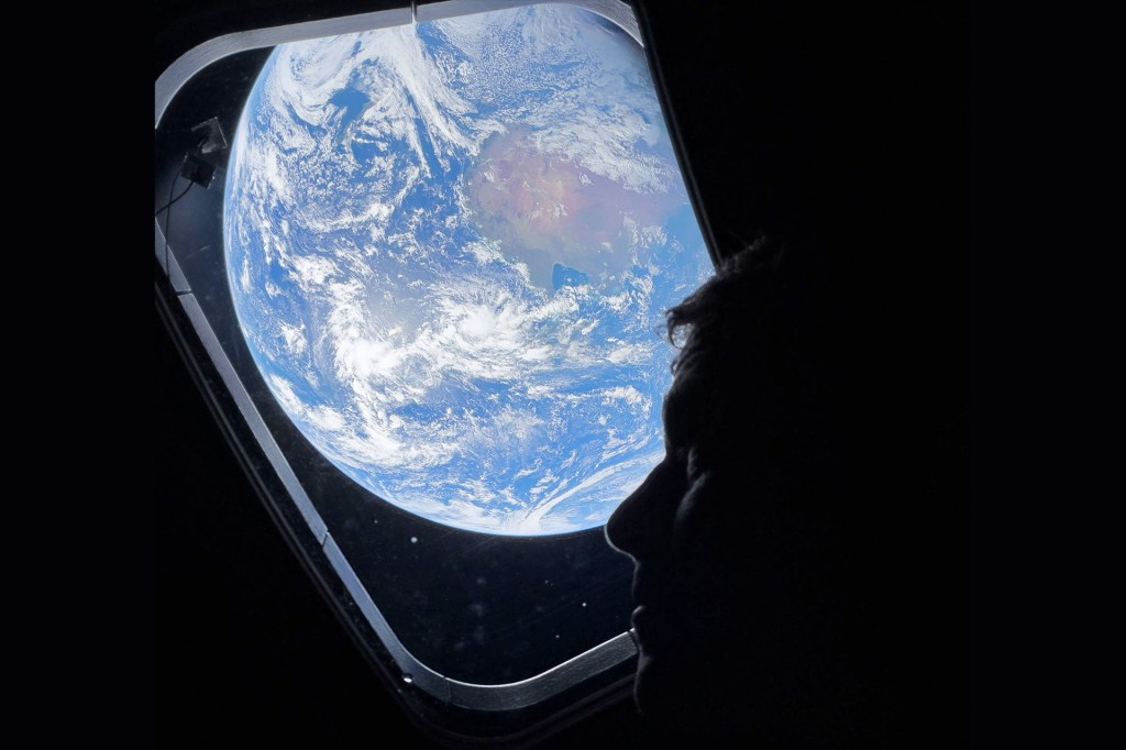 A person looks out a spacecraft window at Earth, which appears bright and blue against the dark background of space.