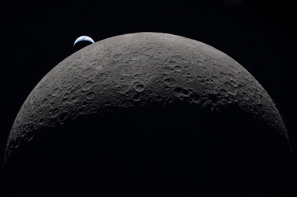 Close view of the Moon’s cratered surface with a small crescent Earth rising in the dark sky behind it.