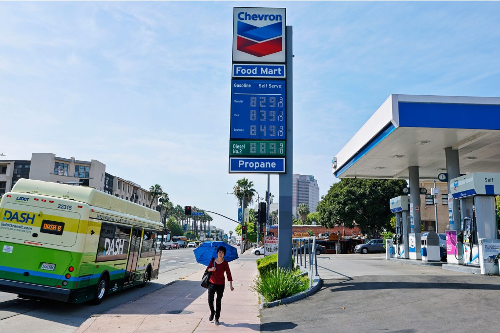A gas station sign shows fuel prices as a bus and a person with an umbrella pass by on the street.