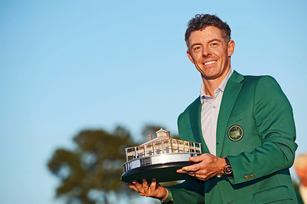 A man in a green jacket smiles while holding a trophy shaped like a building outdoors.