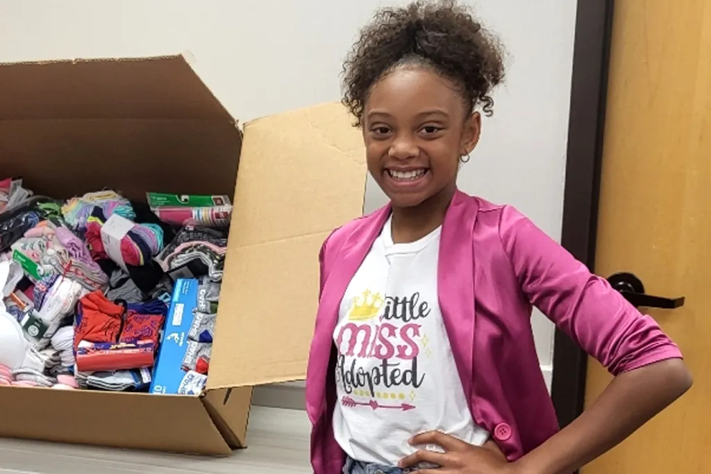 A smiling girl stands next to a large box filled with socks and other donated items.