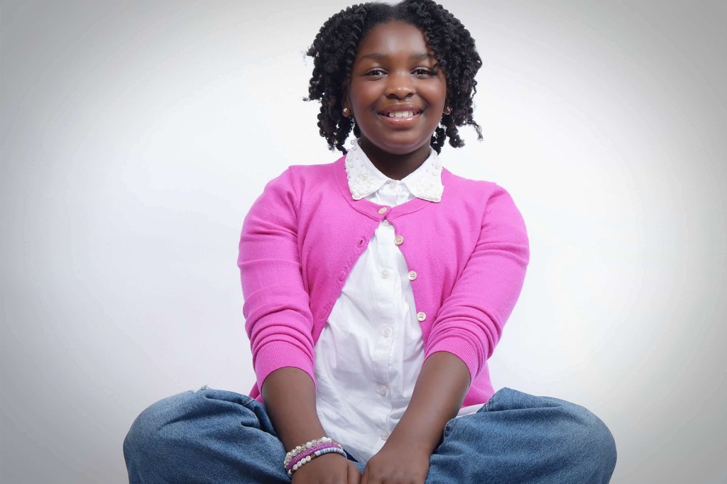 A smiling girl sits cross-legged on the floor, wearing a pink sweater and white shirt against a plain background.