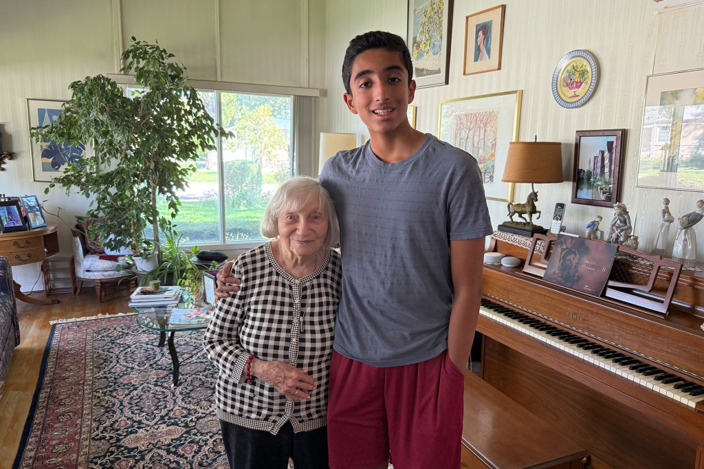 A teenage boy stands beside an elderly woman in a cozy living room, with a piano, plants, and framed pictures behind them.