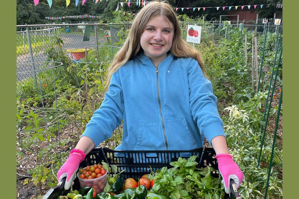 A girl in a blue hoodie and pink gloves holds a crate of fresh vegetables in a garden.