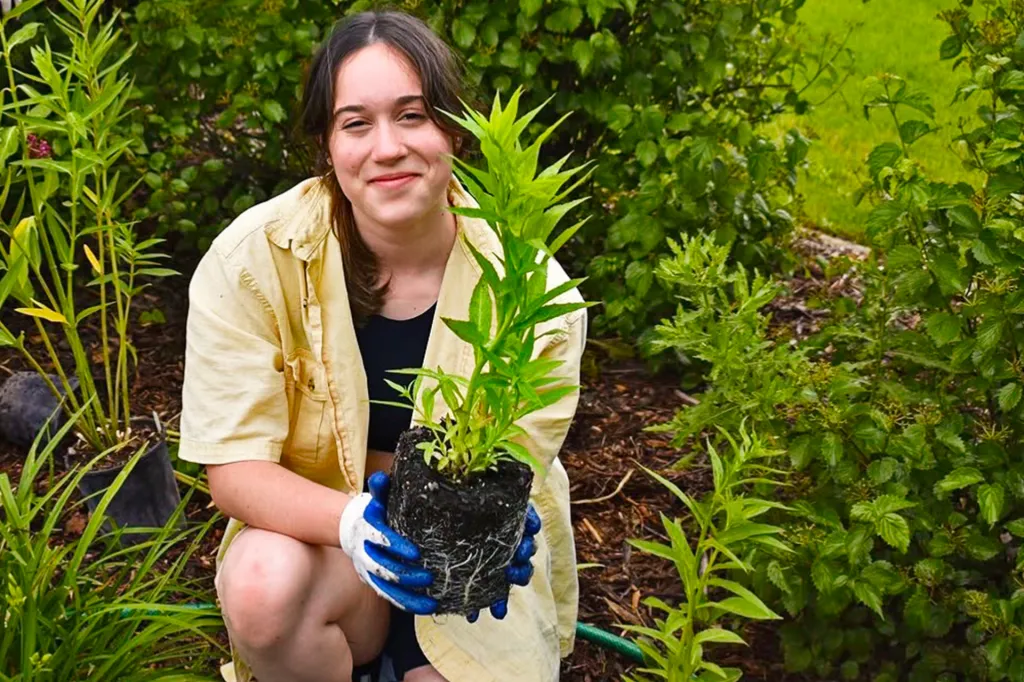 A girl kneels in a garden, smiling and holding a young plant with roots.
