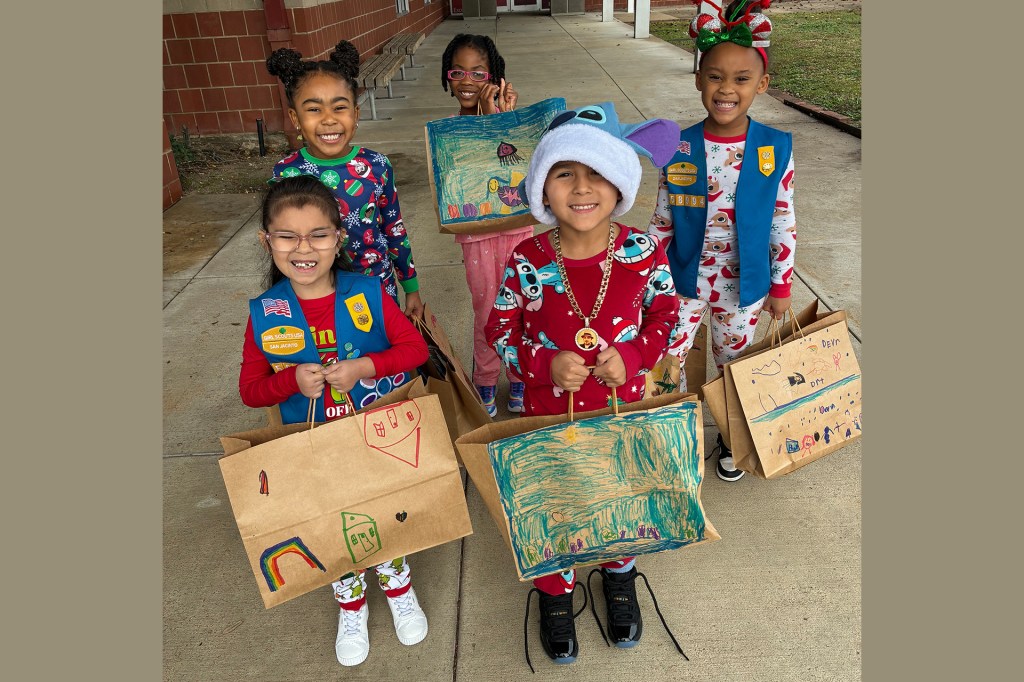 Four young children smile and hold decorated paper bags with drawings while standing outside.