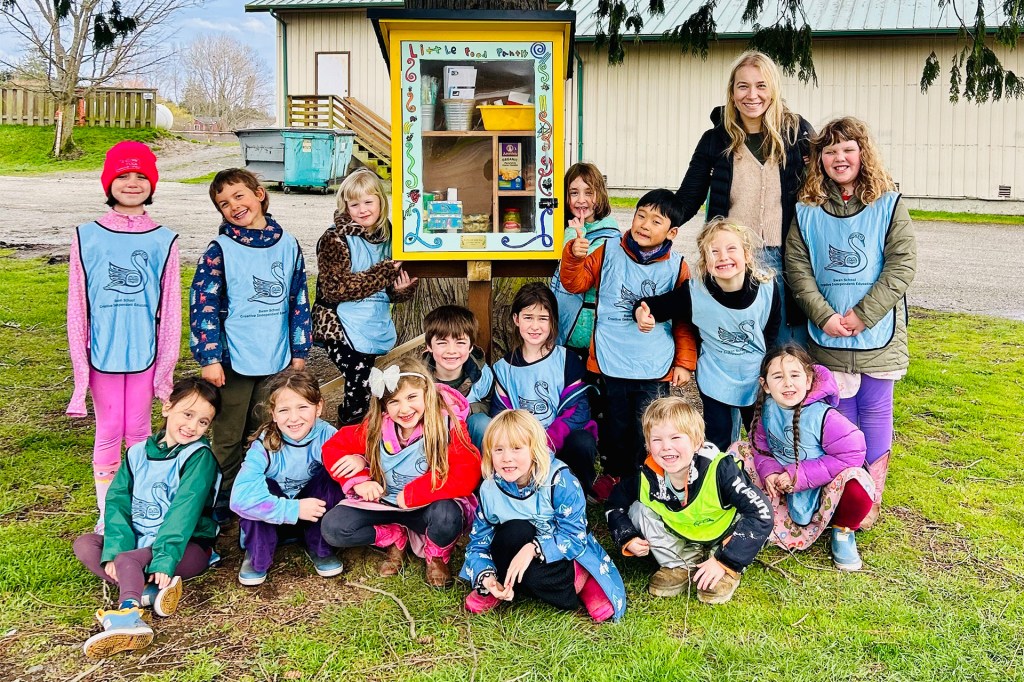 A group of young children and a teacher pose together outdoors around a small food-sharing box.