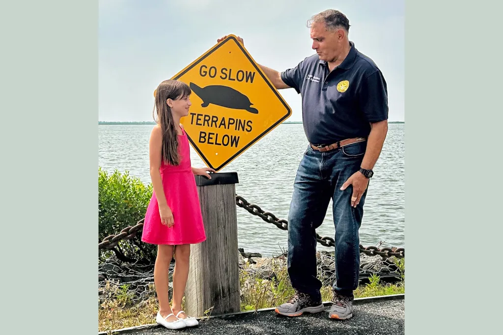 A man and a girl stand by the water next to a yellow road sign that reads “Go slow—terrapins below.”