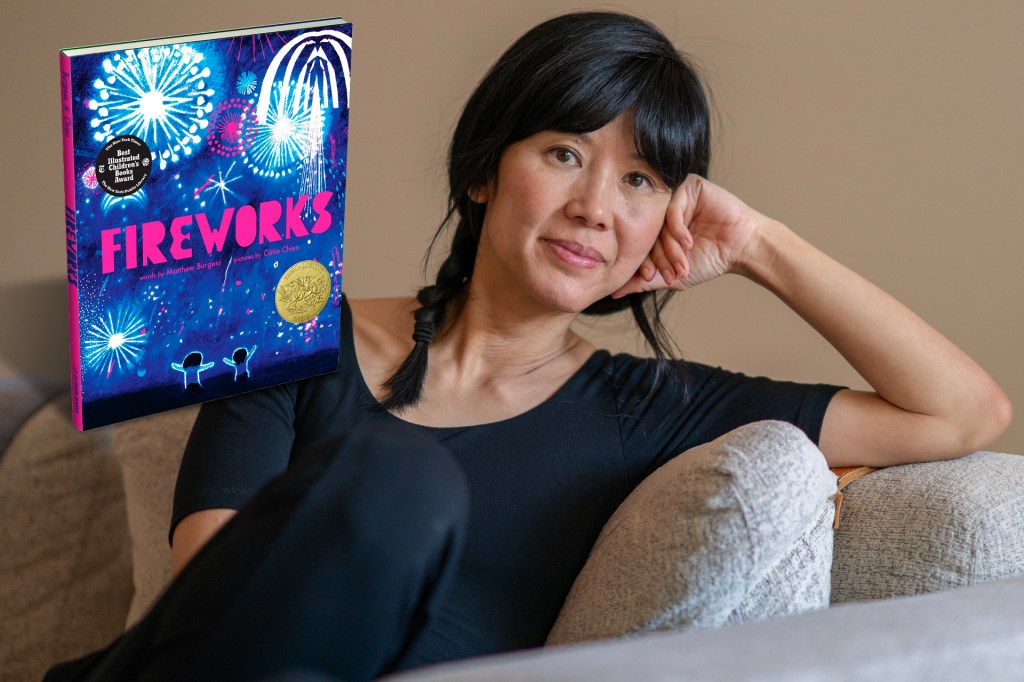 A woman with dark hair sits on a couch, resting her head on her hand, next to a book titled “Fireworks” showing bright fireworks in the night sky.