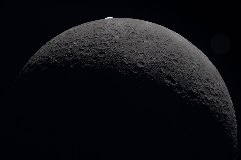 Close-up view of the moon’s crater-covered surface with a small crescent of Earth rising above the horizon against black space.