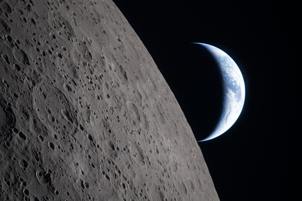 The moon’s gray, cratered surface fills the foreground while a bright crescent Earth glows in the distance against dark space.