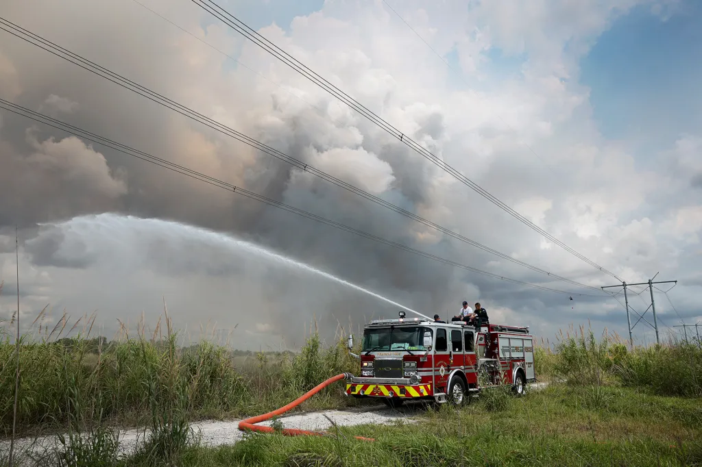 Firefighters spray water from a fire truck into smoky grassland under cloudy skies.