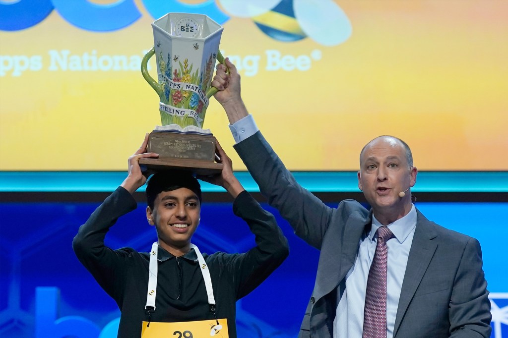 A smiling spelling bee champion holds a large trophy above his head on stage as an event host raises the trophy during the award ceremony.