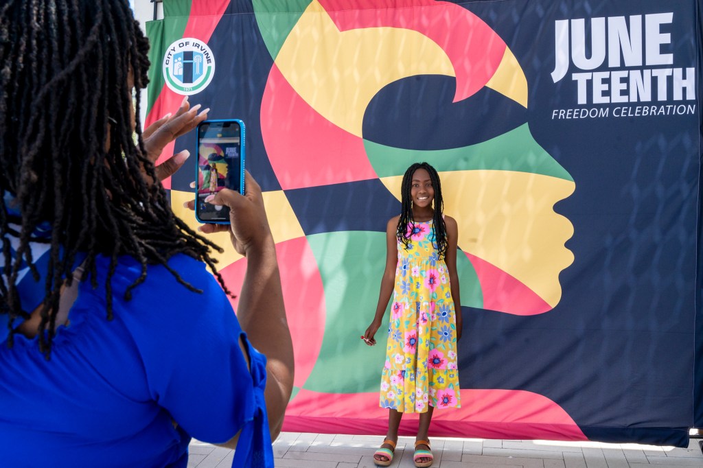 A girl in a colorful floral dress poses in front of a Juneteenth Freedom Celebration banner while an adult takes her photo with a smartphone.
