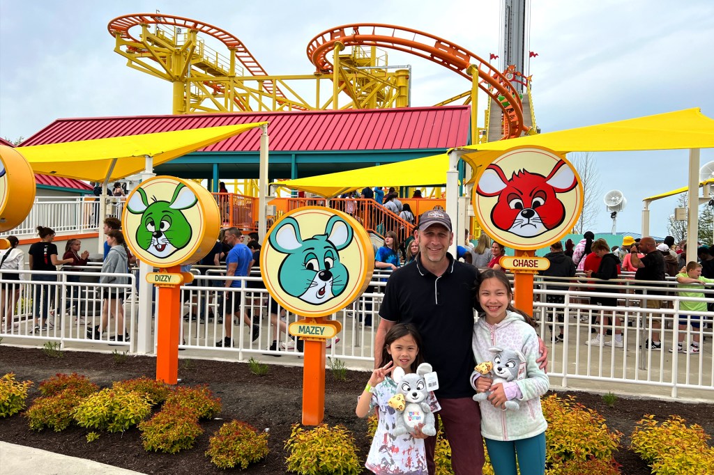 A man and two girls stand in front of a roller coaster at an amusement park.