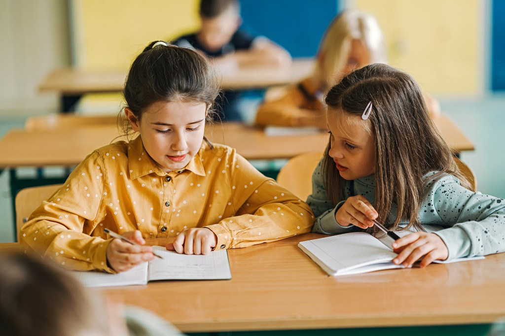 One student writes in a notebook while a classmate looks on.