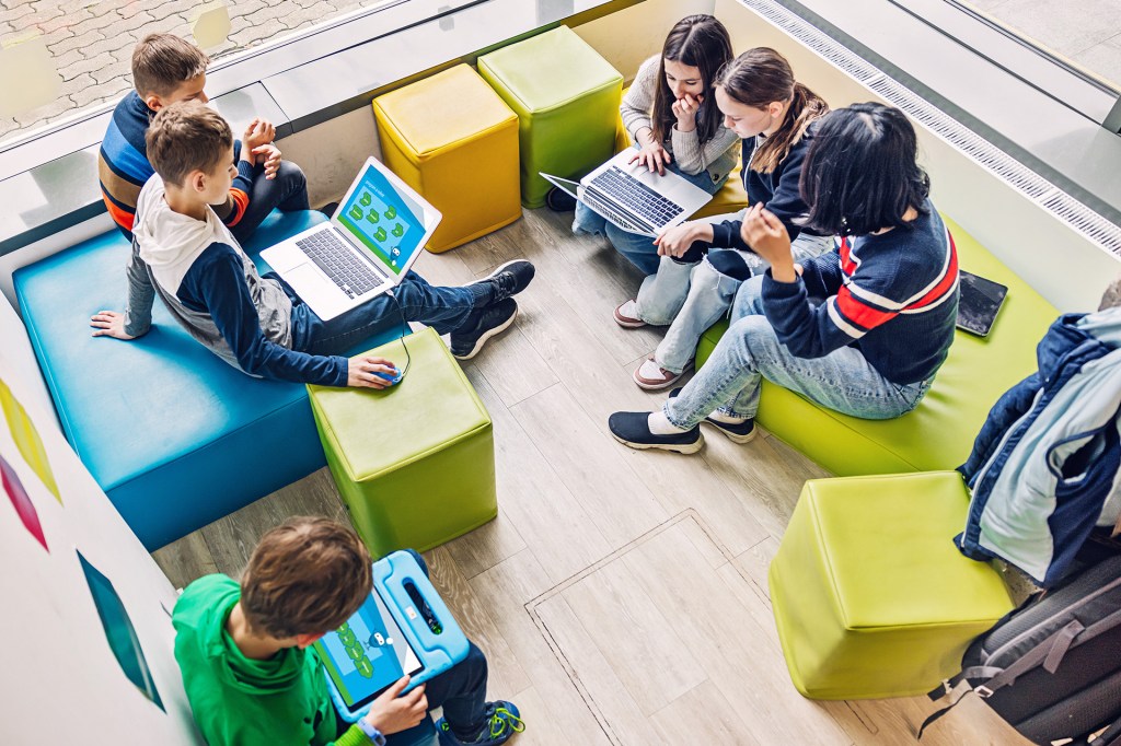 A group of students sitting on colorful seats, working on laptops and tablets.