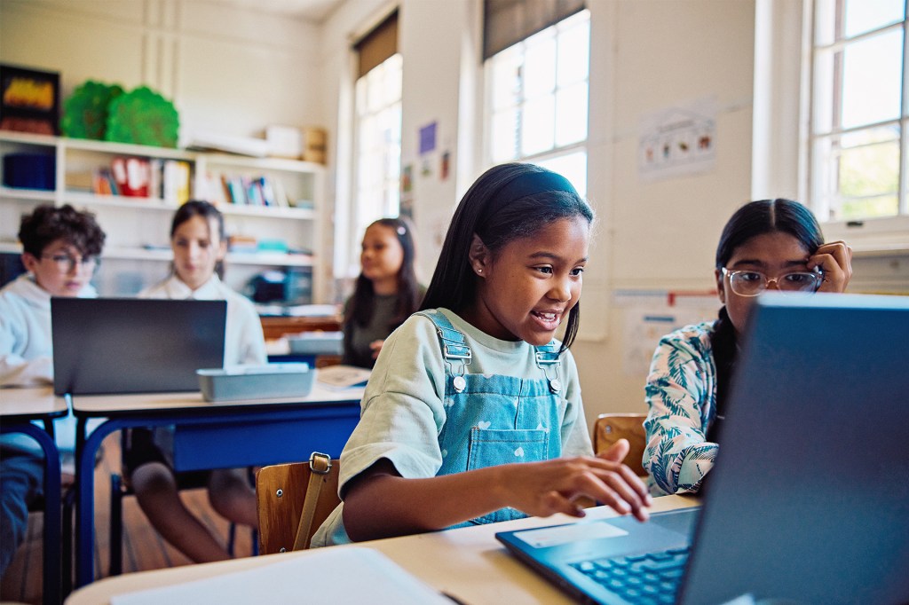 Two students sitting together at a desk look at a laptop, with classmates in the background.