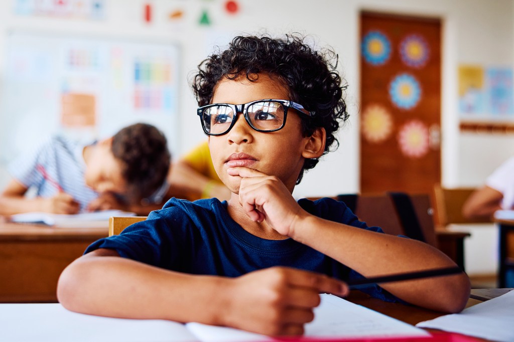 A boy wearing glasses sits at a desk holding a pen with a thoughtful look on his face.