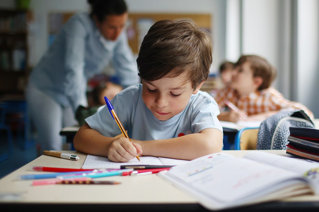 A boy sitting at a desk writing in a notebook. A teacher helps a student in the background.