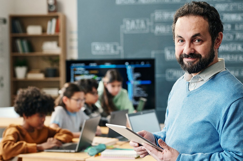 A teacher holding a tablet looks into the camera, and students work on laptops at their desks in the background.