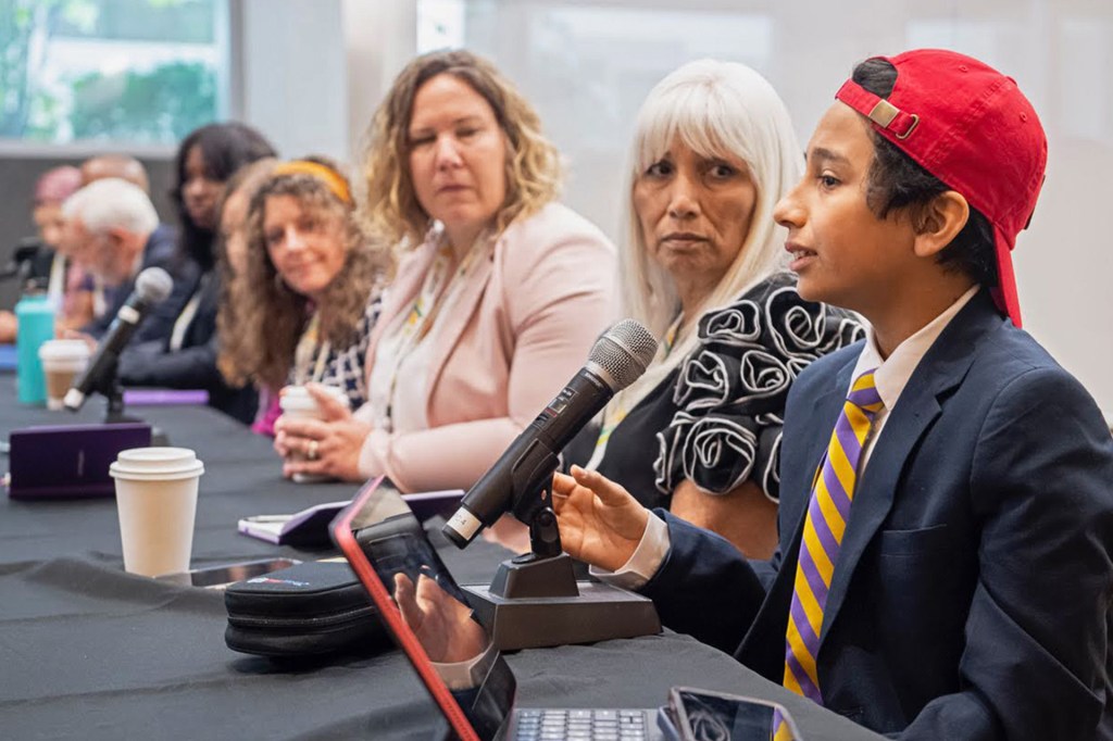 Kid in suit and red cap speaks into a panel microphone.