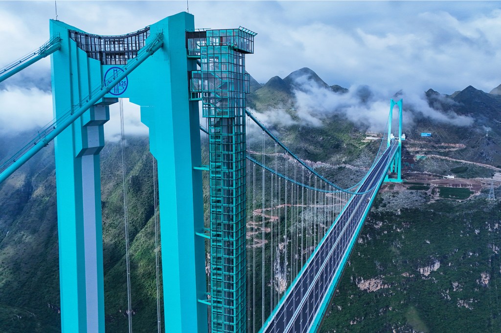 A turquoise suspension bridge spans a deep mountain valley surrounded by clouds.