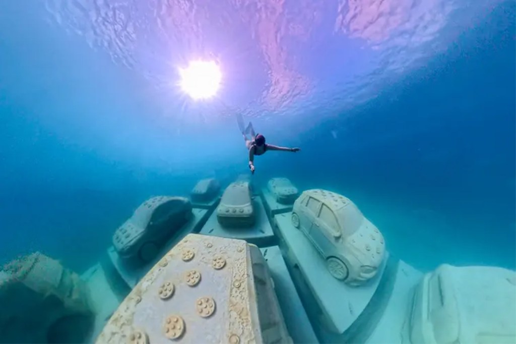 A snorkeler swims above an underwater sculpture of cars on the seafloor.