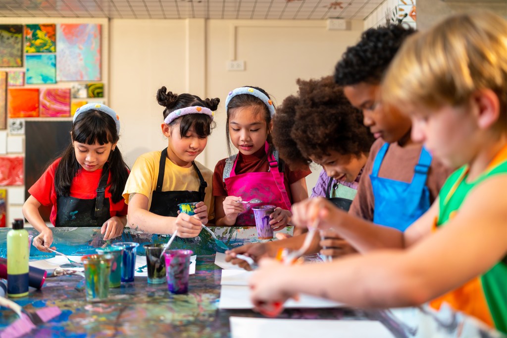 Children wearing aprons work together on colorful art projects at a table covered with paint and supplies.