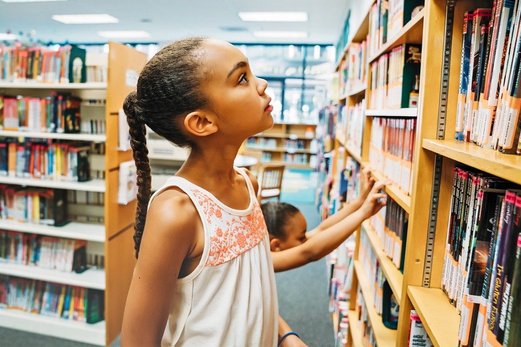 A child looks through books on library shelves while another child reaches for a book nearby.