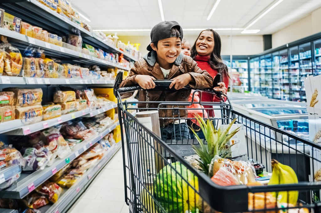 A child smiles while riding in a shopping cart as an adult pushes it through a grocery store.