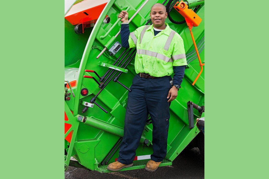 A sanitation worker stands on the back of a bright green garbage truck.