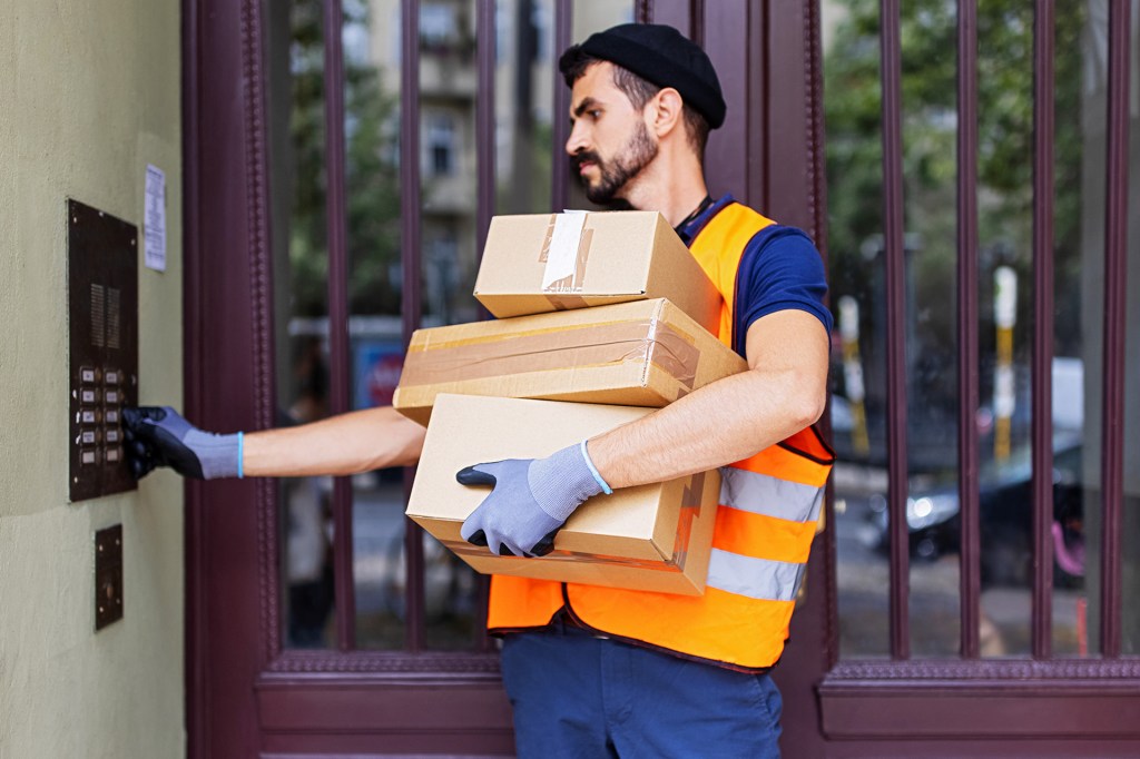 A delivery driver carries boxes while pressing an apartment building buzzer.