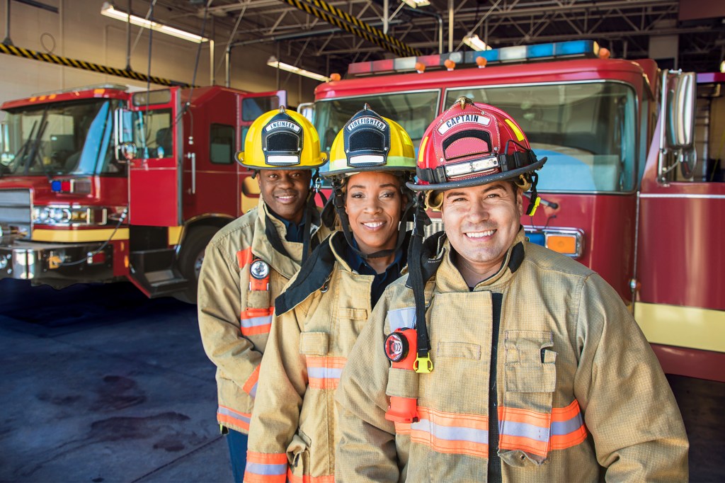 Three firefighters stand smiling in front of fire trucks inside a fire station.