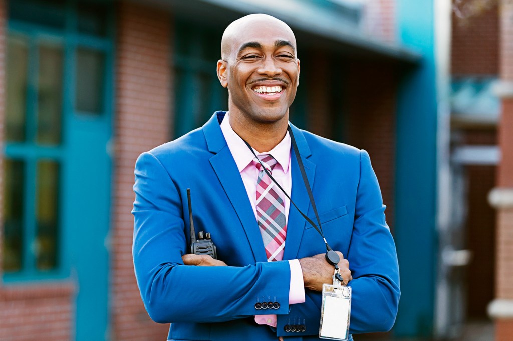 A smiling man wearing a blue suit and carrying a walkie talkie outside a school building.