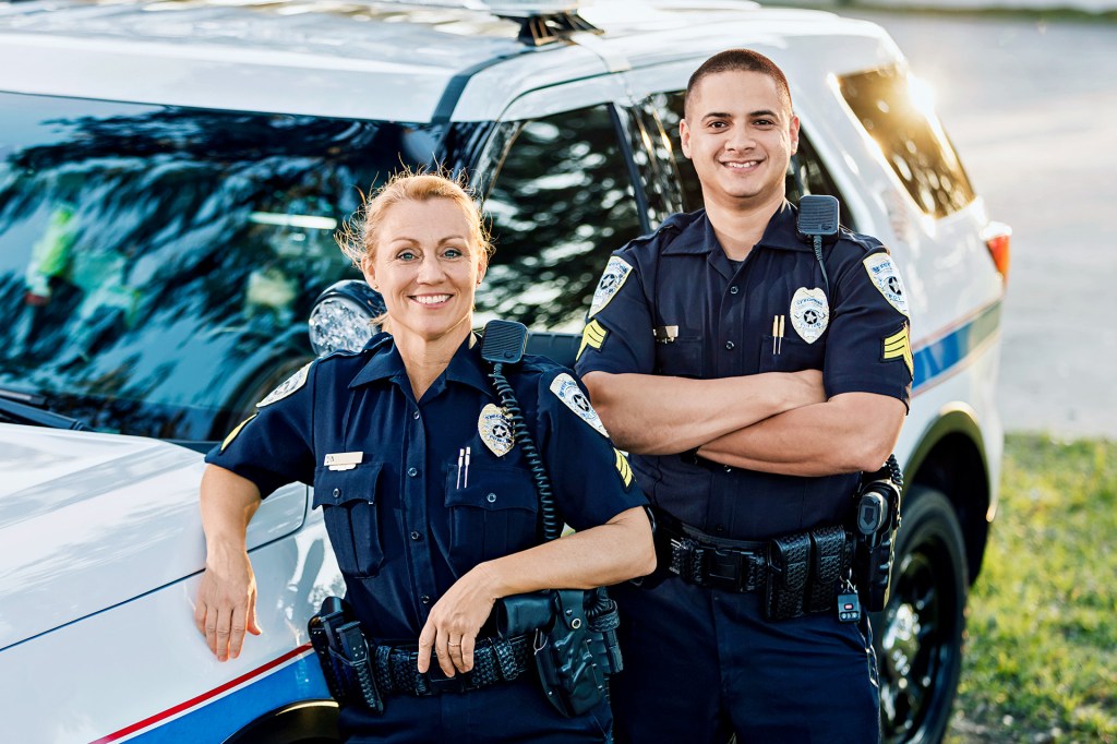 Two police officers stand smiling in front of a police car.