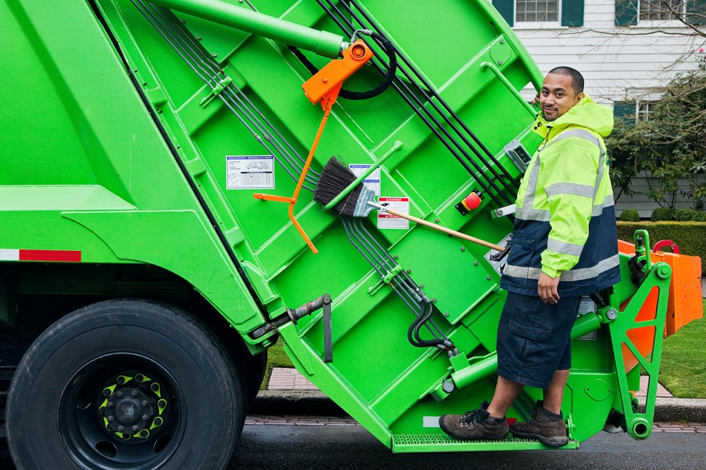 A sanitation worker stands on the back of a bright green garbage truck.