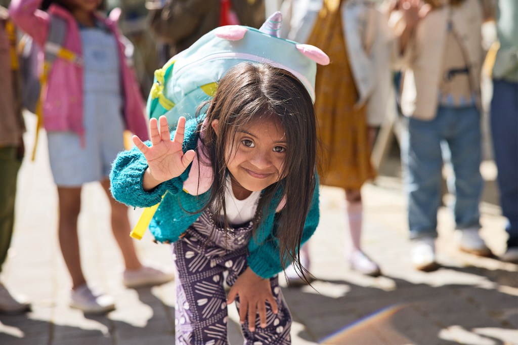 A smiling child waves while wearing a colorful backpack as other children stand nearby.