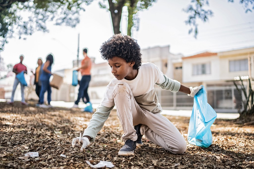 A child wearing gloves picks up litter while holding a blue trash bag.
