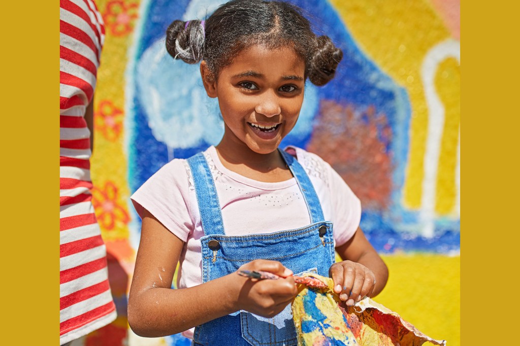 A smiling child holds a paintbrush in front of a bright mural.