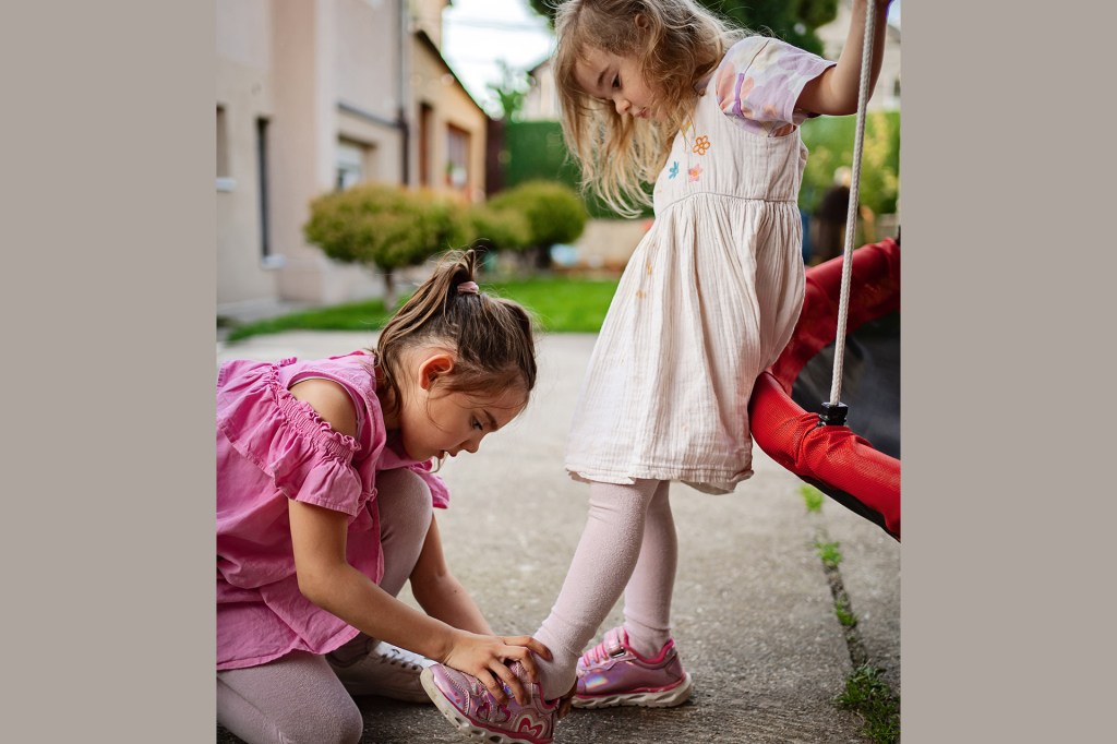 One young child helps another put on a shoe outdoors.
