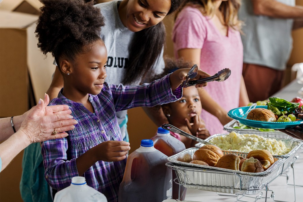A child uses tongs to serve food from a table while adults and other children stand nearby.
