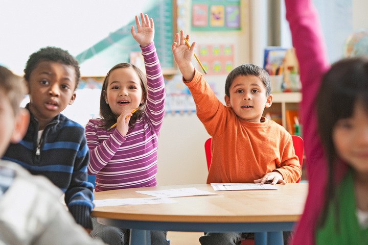 Students sit at desks in a classroom with their hands raised to answer the teacher’s question.